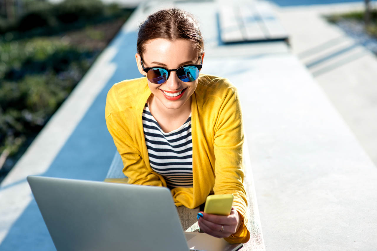 Young woman in yellow sweater working with laptop and phone on the bench in the city