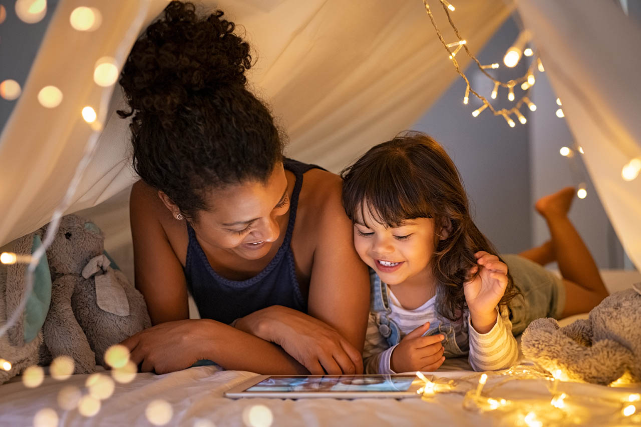African mother and cute smiling girl using digital tablet while lying in illuminated tent in kid bedroom.