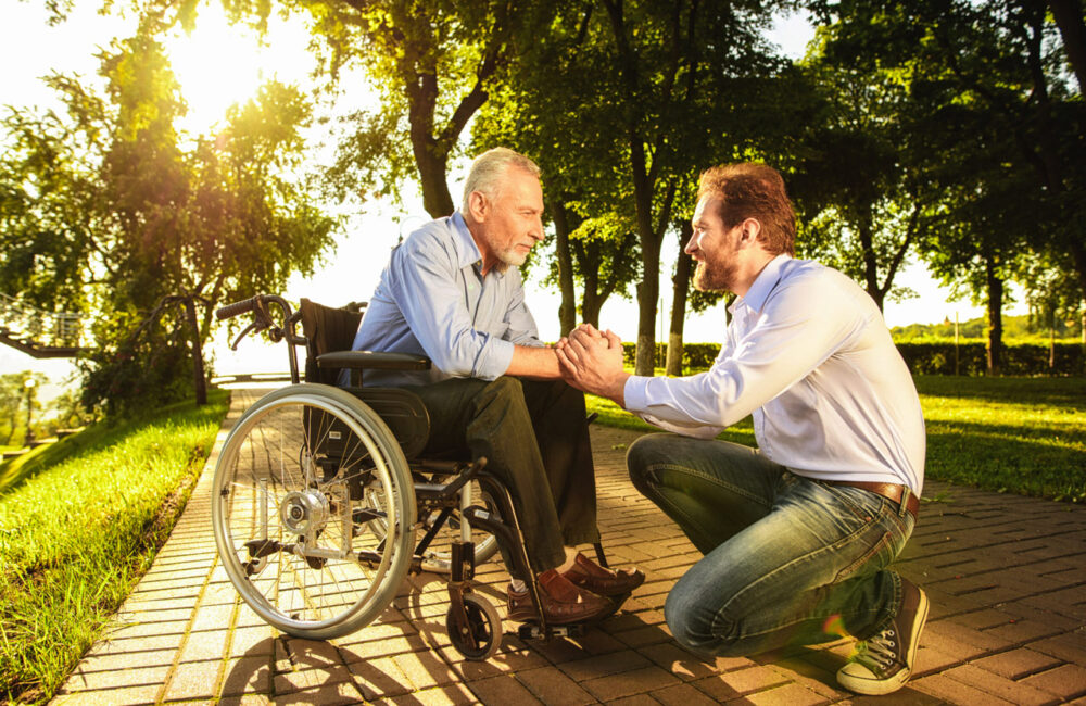 An older man in a wheelchair grasping hands with a younger man crouched in from of the wheelchair