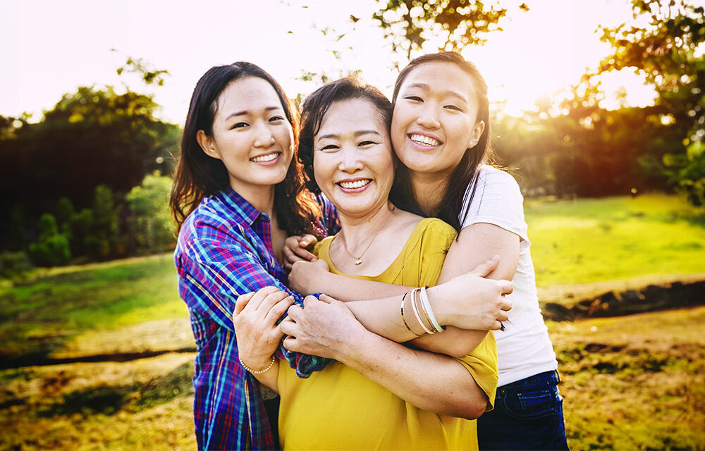 A mother and her two adult daughters hugging in a park and smiling at the camera