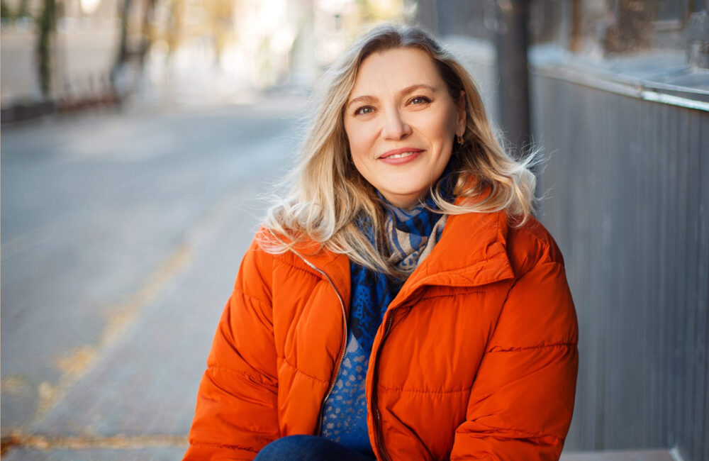 Happy smiling middle aged woman in orange down jacket sitting on concrete stairs outdoors