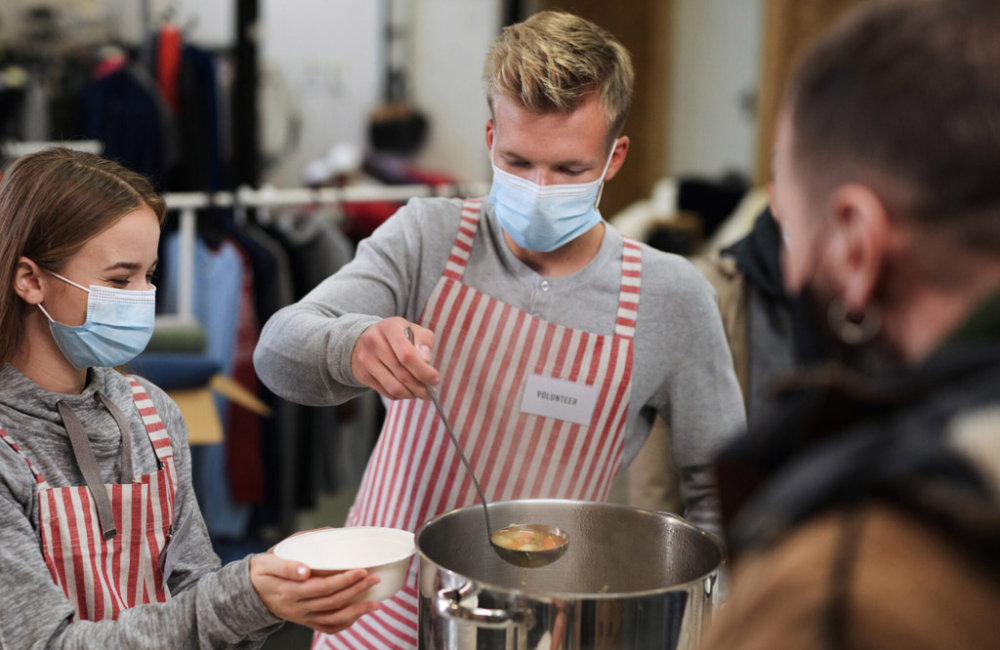 Masked volunteers serving hot soup for homeless in community charity donation center